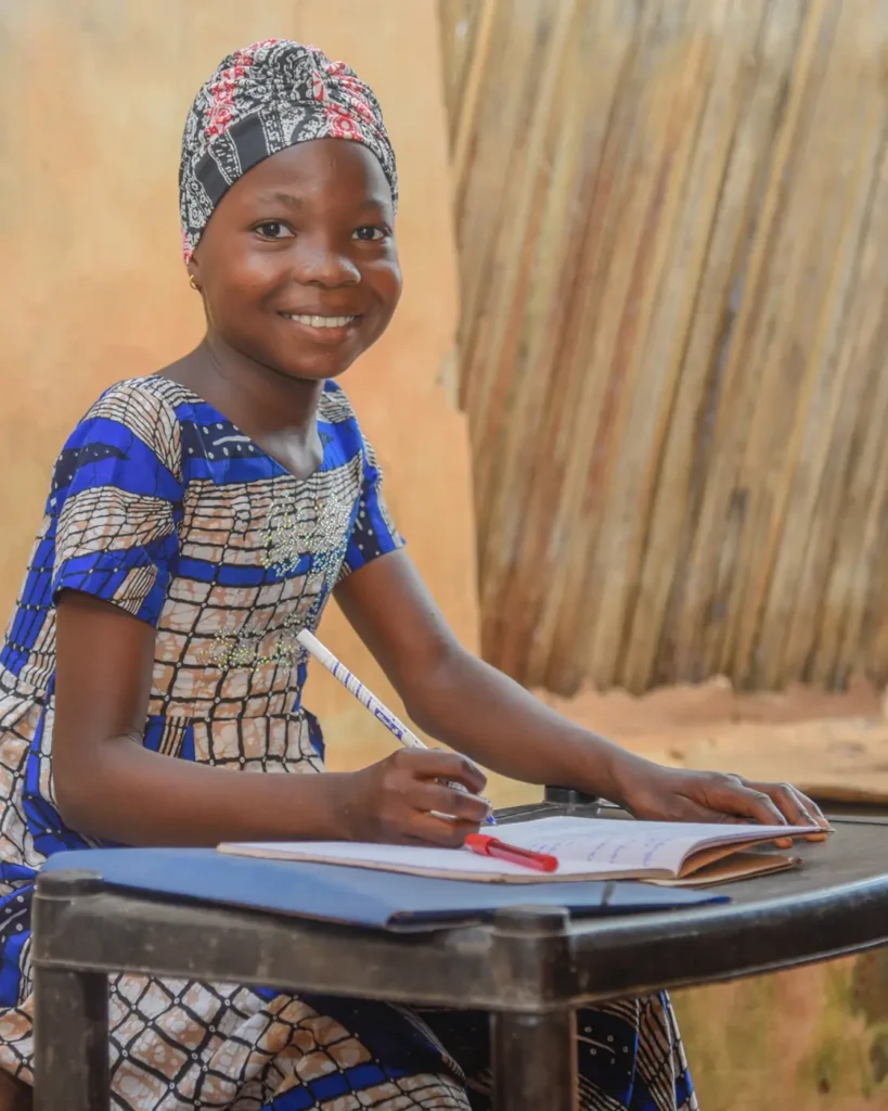 A girl in a patterned headwrap and a blue patterned dress sits at a desk writing in a notebook, smiling at the camera.