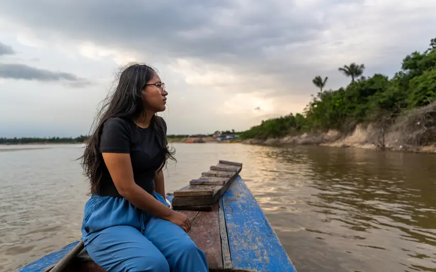 A girl in glasses sits in the bow of a blue boat on a wide brown river, looking out towards the far bank.