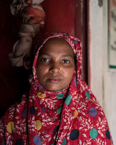 A woman in a red patterned headscarf stands in a doorway, looking steadily at the camera.