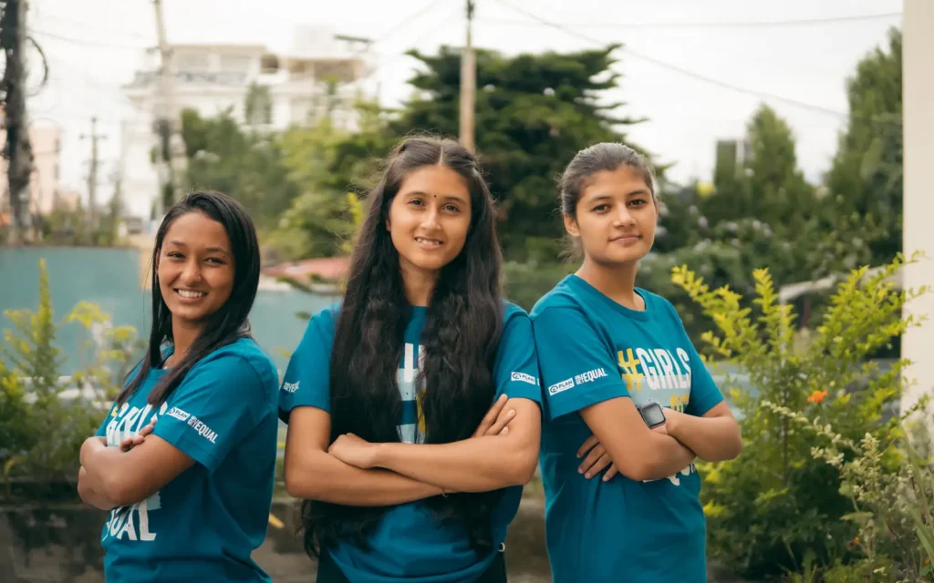 Three young women in teal Girls Get Equal T-shirts stand side by side with their arms folded, smiling.