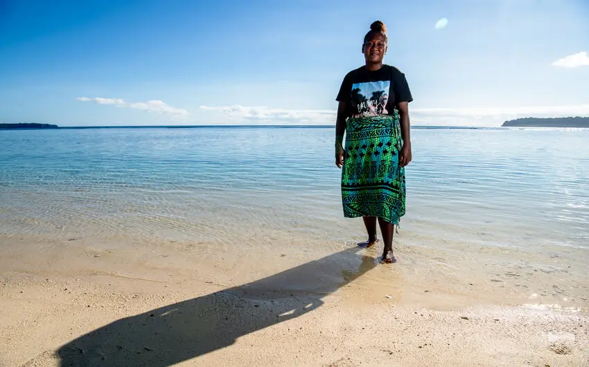 A young woman stands barefoot in clear shallow water at the edge of a beach, under a wide blue sky.