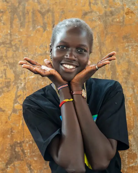 A young woman smiles, framing her face with her open hands, in front of an ochre wall.