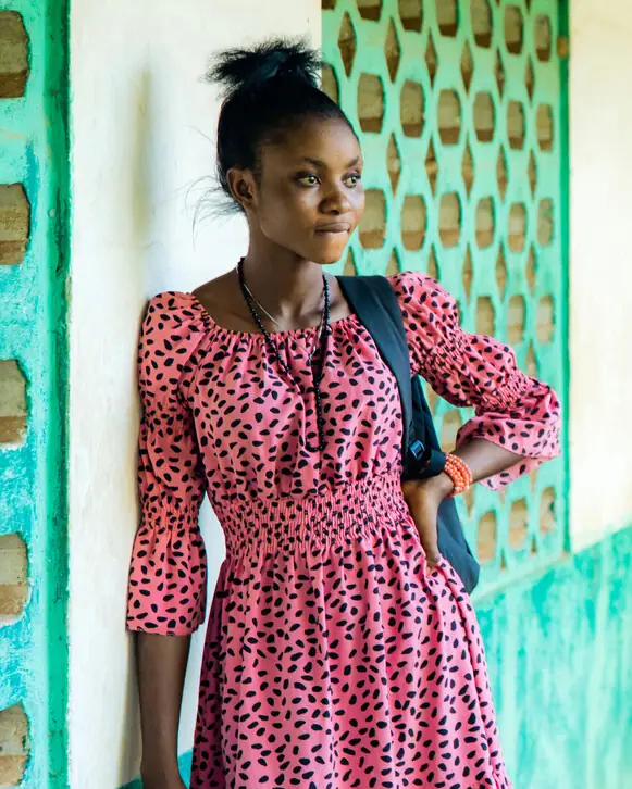 A young woman in a pink patterned dress leans against a green wall, a bag over her shoulder.