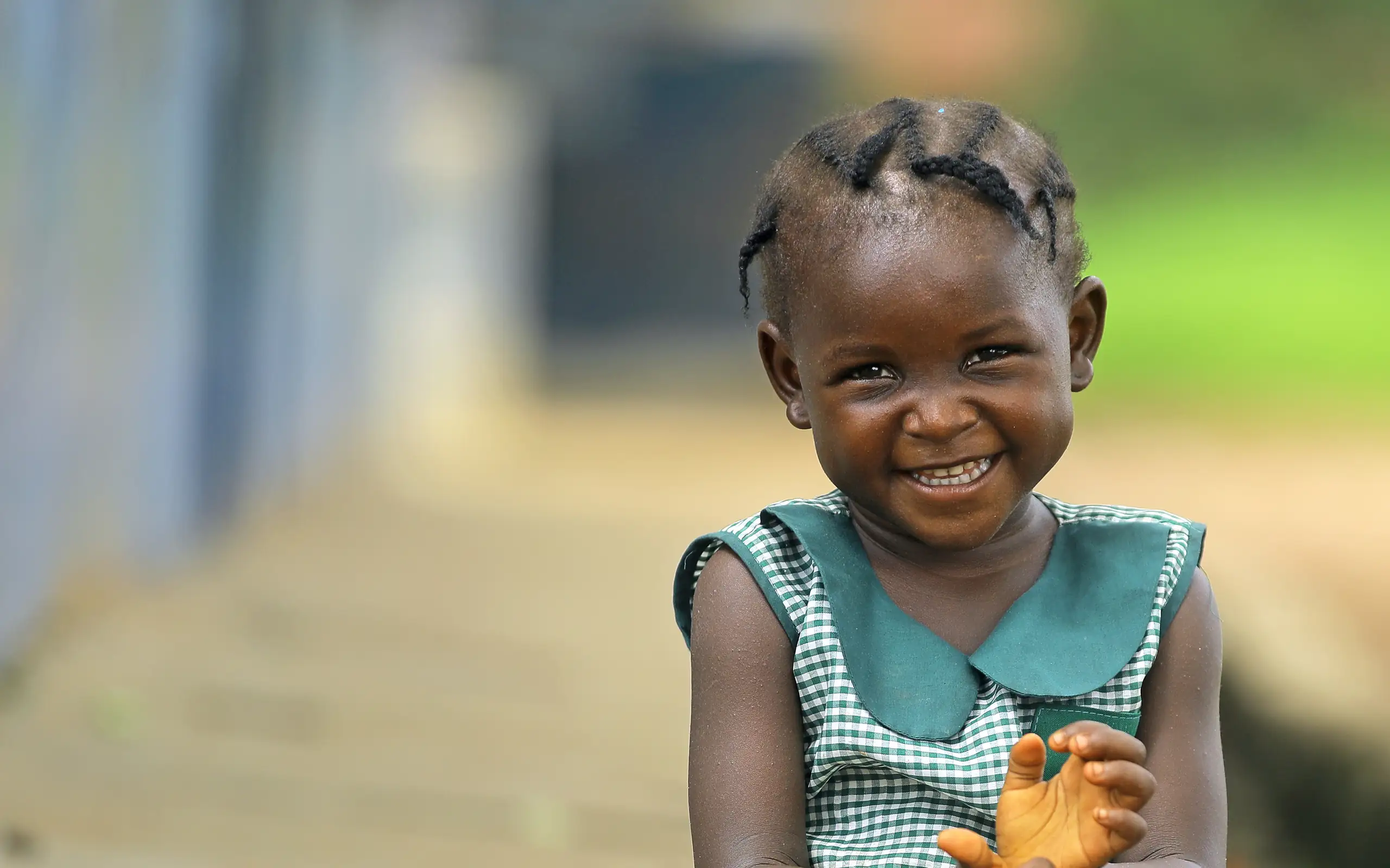 A young girl in a green and white checked school dress sits outdoors, smiling, with her hands together.
