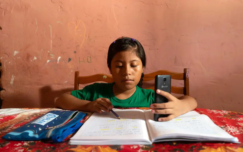 A girl in a green T-shirt writes in an exercise book, holding a mobile phone.