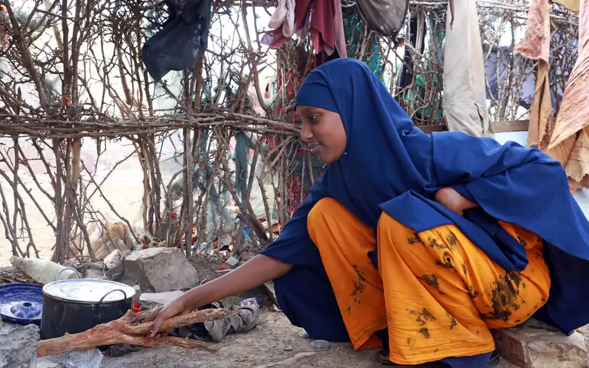 A girl in a blue headscarf crouches beside a cooking pot, feeding the fire.