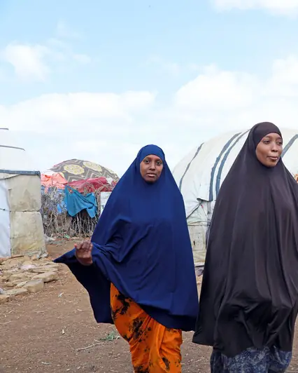 Two girls in long headscarves walk together past shelters.