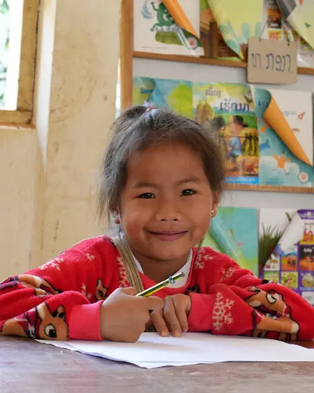 A young girl at her desk with a pencil and paper, smiling at the camera.