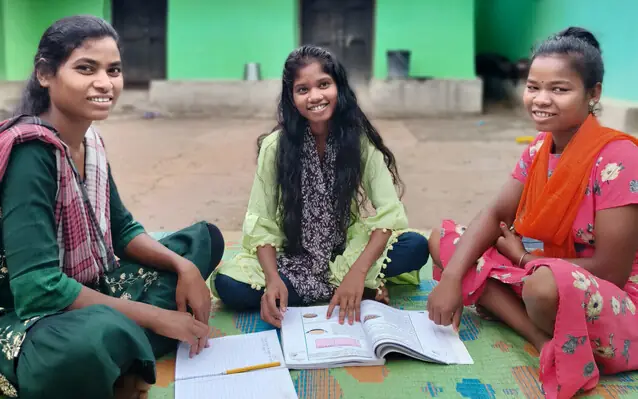 Three girls sit on a mat outside a house, studying with an exercise book and a textbook open, smiling.