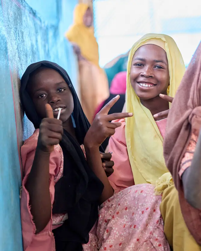 Two girls in headscarves smile at the camera, one giving a thumbs-up, the other a peace sign.
