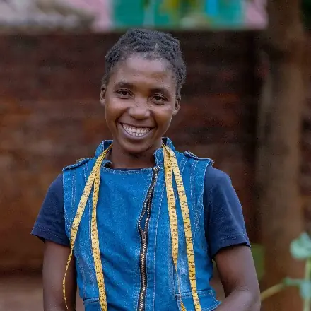 A young woman in a denim waistcoat, a yellow tape measure around her neck, smiles at the camera.