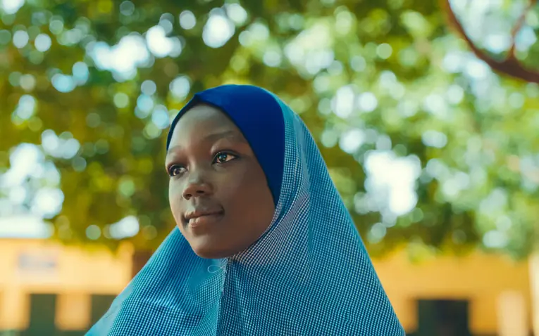 A girl in a blue checked headscarf looks off to one side, leafy green trees softly blurred behind her.