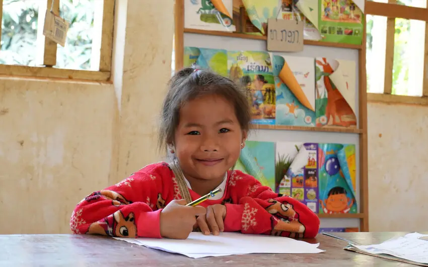 A young girl at her desk with a pencil and paper, smiling at the camera.