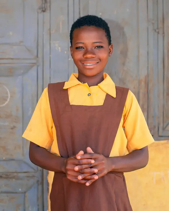 A schoolgirl in a yellow shirt and brown pinafore stands in front of a door, smiling.