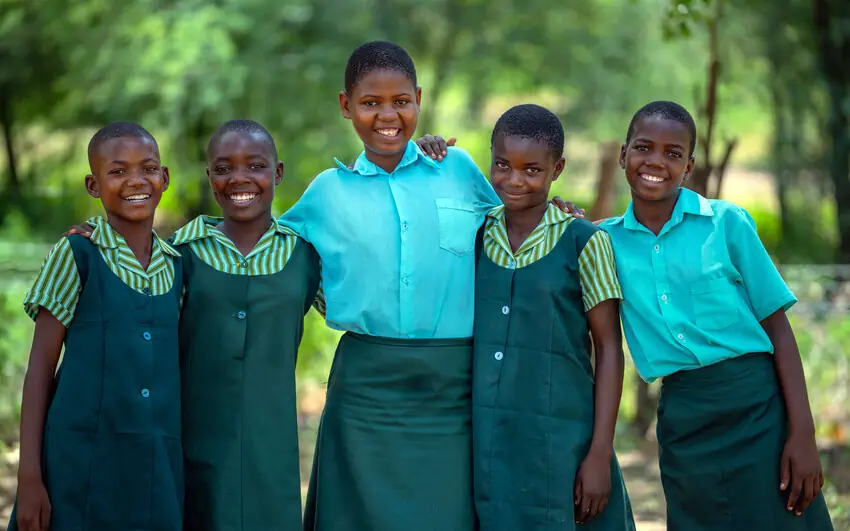 Five schoolgirls in uniform stand with their arms around each other’s shoulders, smiling.