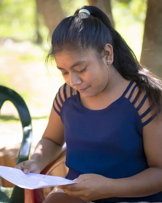 A teenage girl seated outdoors on a plastic chair, reading a printed handout.
