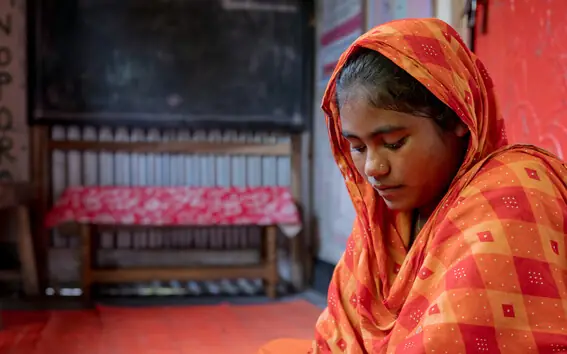 A teenage girl in an orange headscarf looks down at her work in a learning centre, a blackboard behind her.