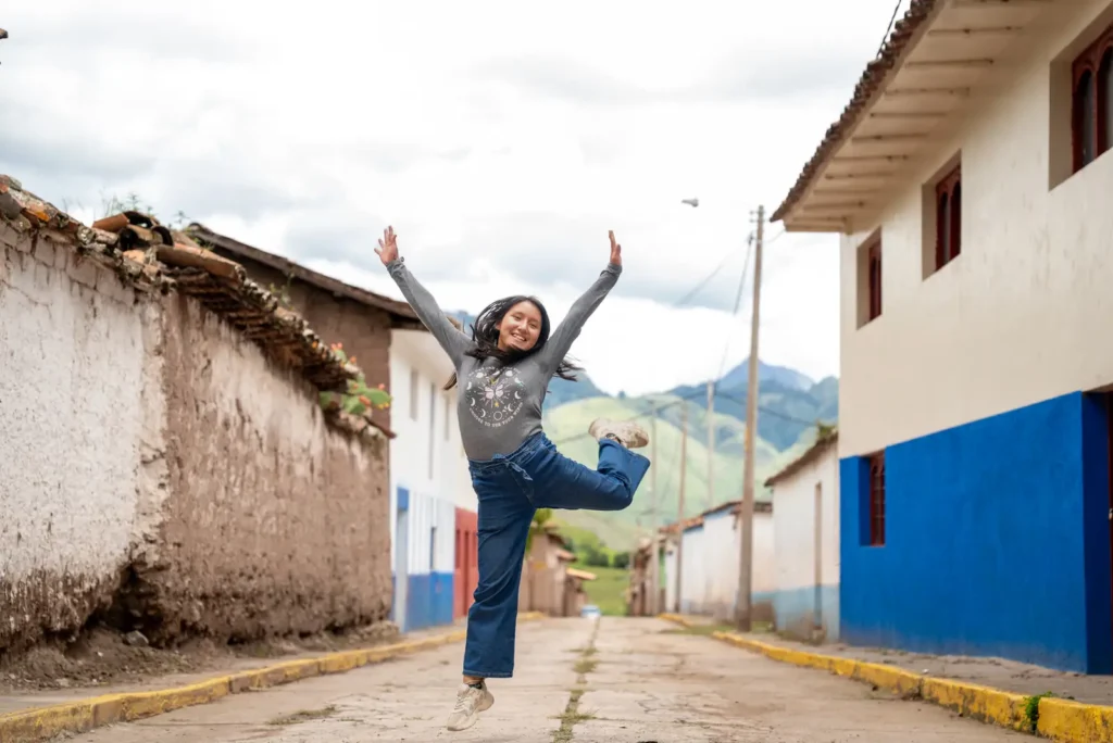 A girl leaps into the air with her arms raised in the street of a hillside village.
