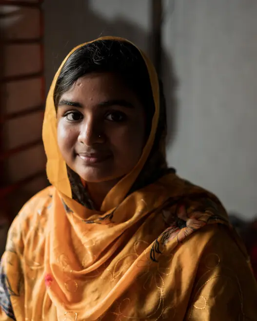 A teenage girl in an orange headscarf looks into the camera with a slight smile.