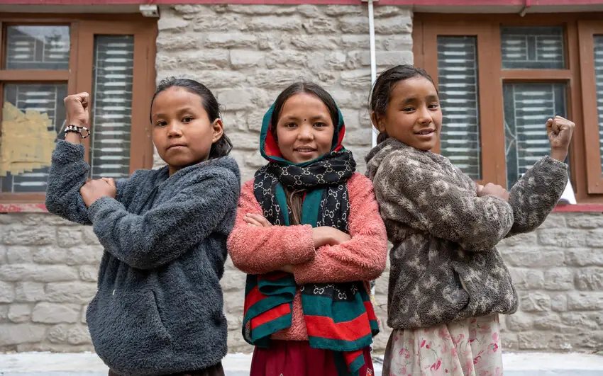 Three girls stand side by side outside a stone building, flexing their arms.