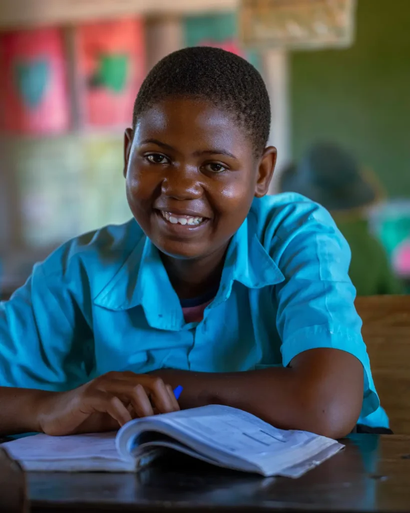 A schoolgirl in a blue shirt sits at a desk in a classroom, her hand on an open book, smiling at the camera.